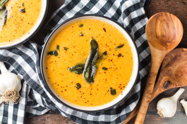 Overhead shot of a bowl of butternut squash soup with sage, wooden spoons, black and white checkered kitchen towel.