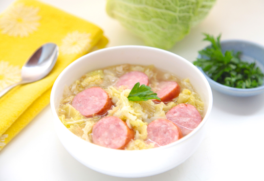 White bowl of cabbage soup with sausage. White background, head of green cabbage.