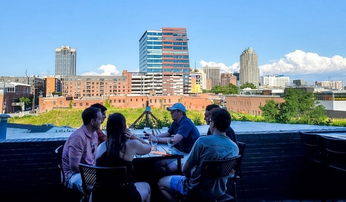 A group of people sit at a table at Wye Hill Kitchen & Brewing with the skyline of Raleigh, North Carolina in the background