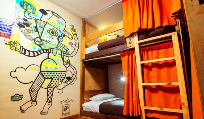 A vibrant mural on the wall next to wooden dorm beds with red curtains at Viajero Kokopelli, a hostel in Cusco, Peru
