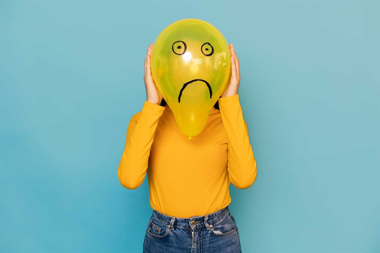 Young woman hidden behing a balloon with a sad face drawn on it over blue background. Negative emotion concept