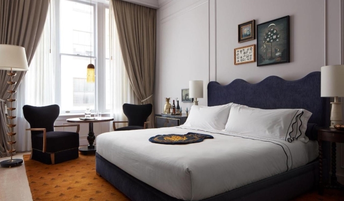 A luxurious guest room with a king sized bed, framed artwork on the walls, and a small round table with easy chairs in front of a large window framed with draperies at Maison Métier, a five-star hotel in New Orleans, Louisiana