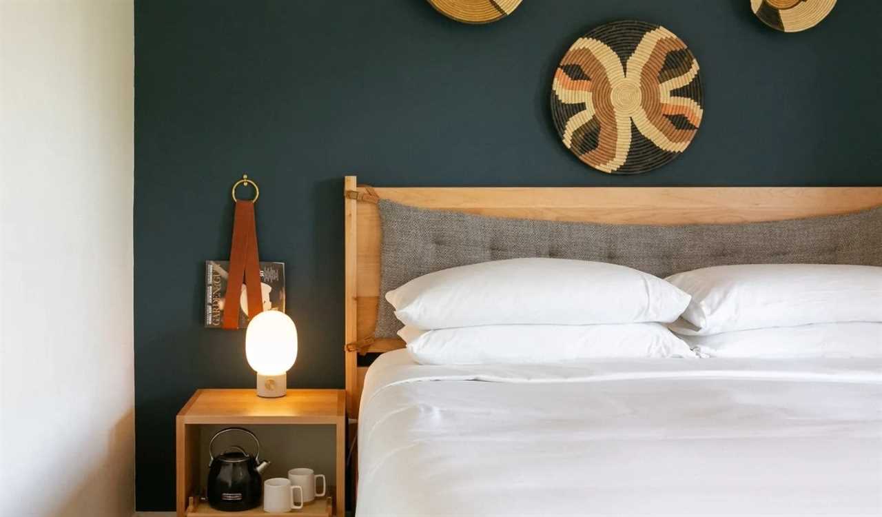 Close up of a wooden nightstand next to a bed with crisp white sheets, against the dark blue walls of Bode, an apart-hotel in Nashville, TN