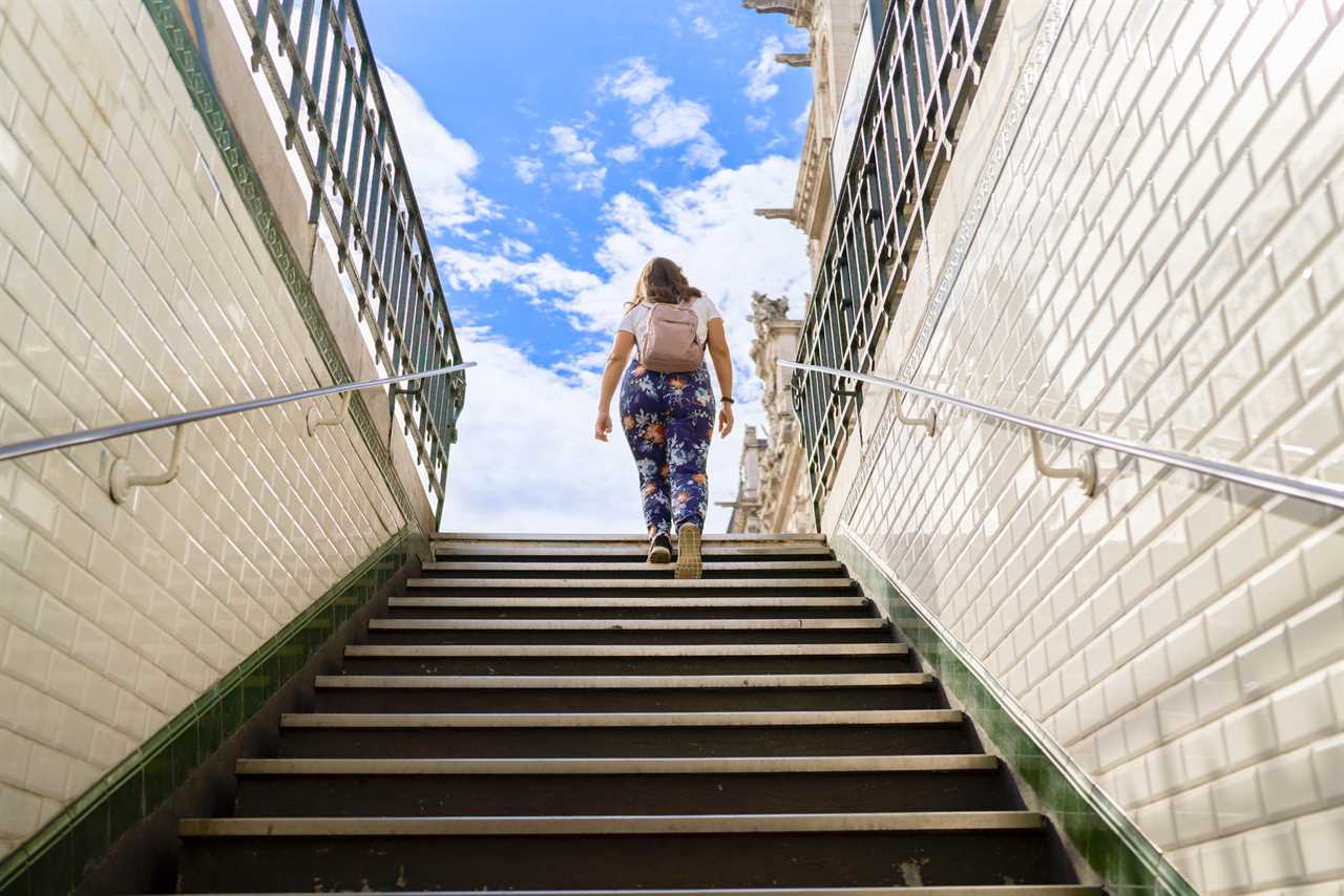 brown-haired Caucasian woman wearing casual clothes with patterned pants, walking up the stairs of the subway, while carrying a backpack, in the background is the partially cloudy sky.