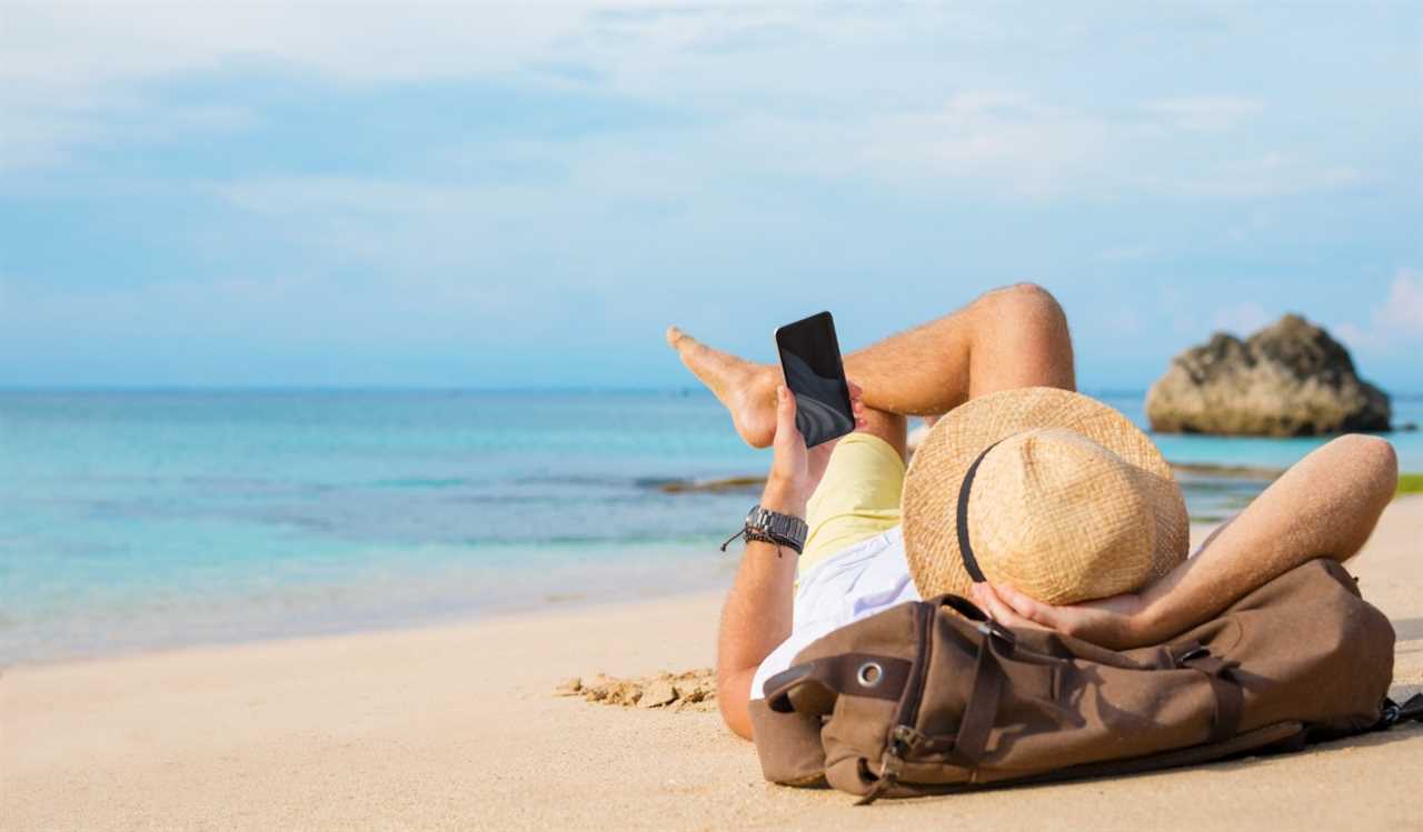 A man in a fedora lying on a beach holding up a smartphone