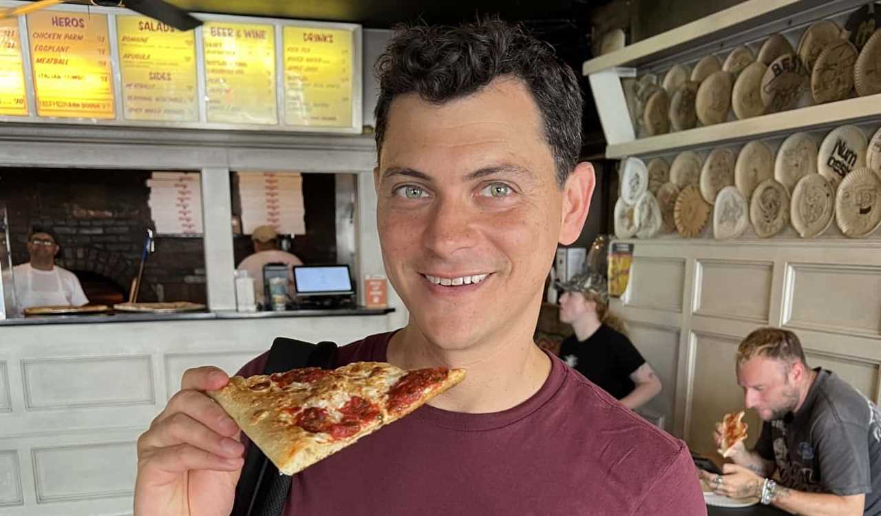 Nomadic Matt holding up a piece of pizza at a pizza shop in NYC